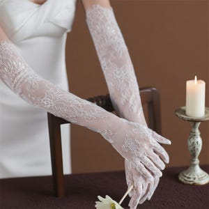 May include: Delicate white lace gloves featuring detailed floral designs. These gloves extend beyond the elbows, offering a refined appearance. Ideal for weddings or special occasions.