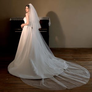Pode incluir: Um vestido de noiva branco com uma longa cauda e um véu a condizer. O vestido tem um design simples e elegante, e o véu cai pelas costas. A noiva está em frente a um piano, com um chão de madeira.