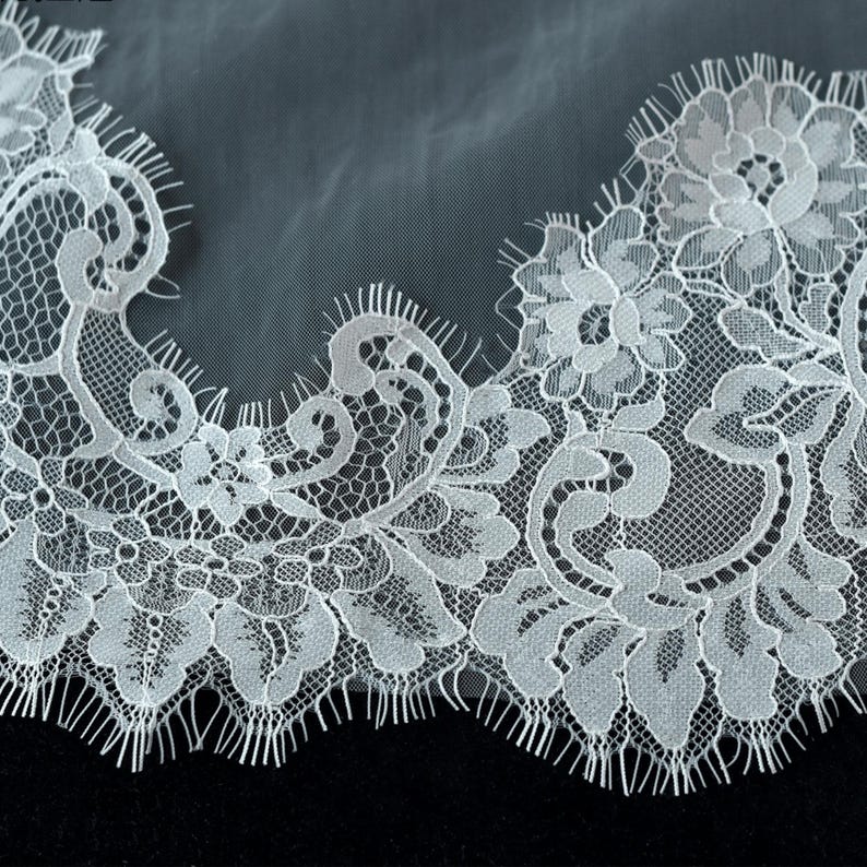 May include: A white lace trim featuring an intricate floral pattern with scalloped edges and fine eyelash fringe details. The delicate fabric displays a complex mesh background with dense embroidery forming petals and leaves along the border.