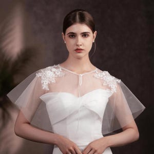 May include: A sheer white bridal capelet with delicate floral lace appliqués on the shoulders. The capelet is made of a soft, flowing fabric and is designed to be worn over a white wedding dress. The capelet adds a touch of elegance and romance to the bridal look.