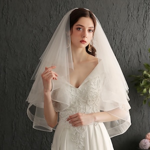 Peut inclure: Une mariée porte une robe de mariée blanche avec un voile en tulle à plusieurs niveaux. La robe présente des détails de dentelle complexes sur le corsage. Le voile est en tissu doux et transparent, créant un look délicat et élégant.