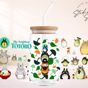 May include: A clear glass tumbler with a wooden lid and a glass straw, decorated with various Totoro character illustrations and green leaves. The tumbler has the text "My Neighbor Totoro" on it.