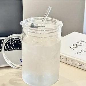 May include: A clear glass mug with a handle, filled with a light-colored liquid, and a clear lid with a straw. The mug sits on a white surface next to a laptop and a book titled "ARTS THE CRAFTS".
