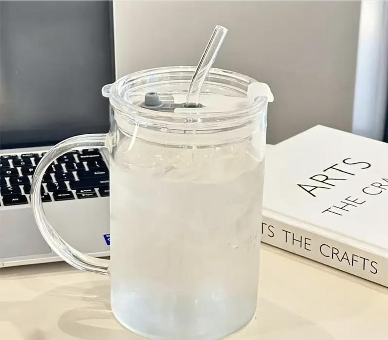 May include: A clear glass mug with a handle, filled with a light-colored liquid, and a clear lid with a straw. The mug sits on a white surface next to a laptop and a book titled "ARTS THE CRAFTS".