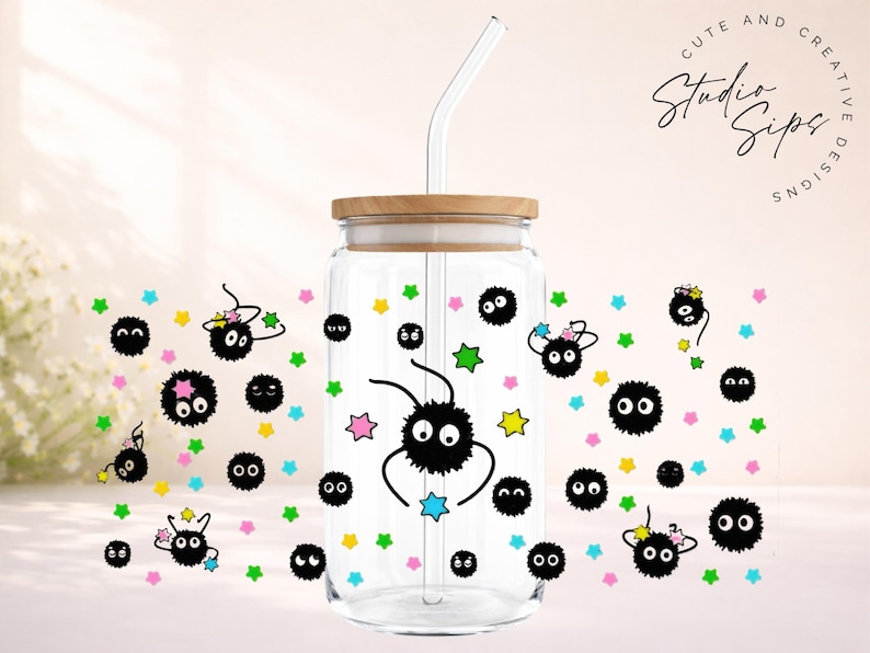 May include: Clear glass tumbler with a wooden lid and a glass straw. The tumbler is decorated with black soot sprites and colorful stars. The text "CUTE AND CREATIVE DESIGNS Studio Sips" is visible on the top right.