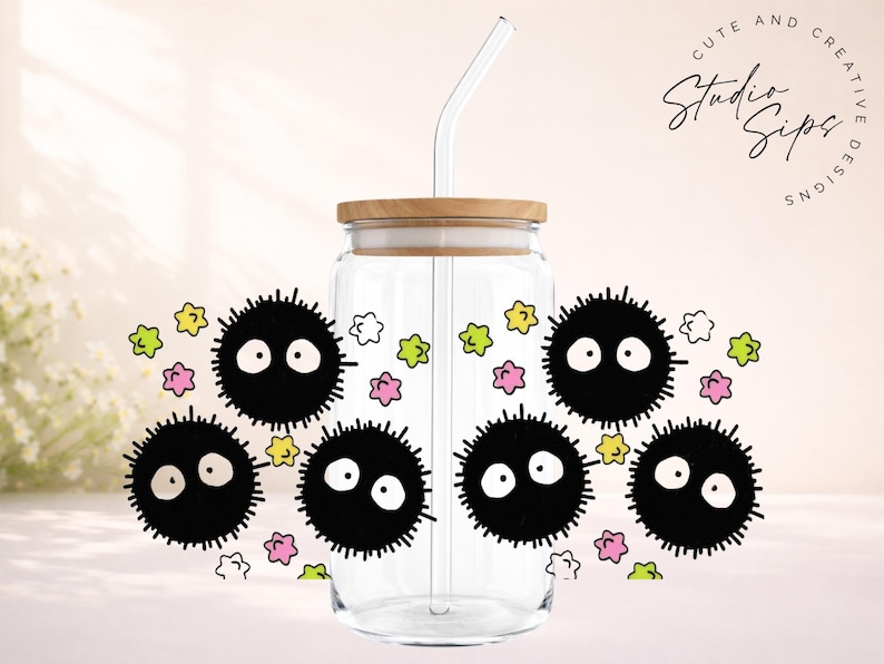 May include: Clear glass tumbler with a bamboo lid and a glass straw. The tumbler is decorated with black soot sprites and colorful star-shaped accents. The text "Cute and Creative Studio Sips Designs" is also visible.