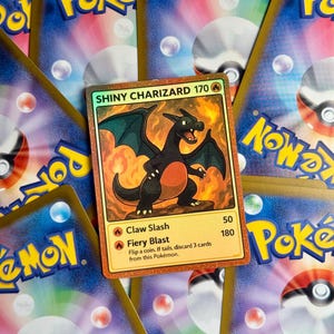 Puede incluir: Una carta coleccionable Pokémon Shiny Charizard con una ilustración de dragón negro, sobre un fondo de otras cartas Pokémon. La carta muestra "SHINY CHARIZARD 170" y detalles de ataque. Las cartas de fondo tienen un diseño azul y amarillo.