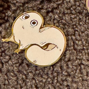 Könnte beinhalten: Ein hellbeige Emaille-Anstecknadel in Form einer stilisierten Kreatur mit großen Augen und kleinen Füßen. Die Anstecknadel hat einen goldenen Umriss und goldene Akzente. Die Anstecknadel befindet sich auf einer strukturierten braunen Oberfläche.