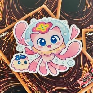 Puede incluir: Una pegatina troquelada con un simpático personaje de dibujos animados rosa con grandes ojos azules, un adorno para la cabeza en forma de flor y un halo azul claro en forma de burbuja. También hay un personaje más pequeño y a juego. La pegatina está colocada sobre un fondo de cartas coleccionables.
