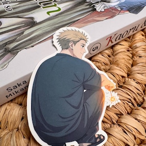May include: A die-cut sticker featuring an anime-style illustration of a man in a dark blue outfit, holding a sparkler. The sticker is placed on a book with Japanese text and a woven surface.