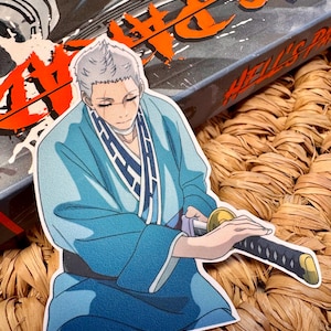 Puede incluir: Una pegatina con un personaje de anime con un kimono azul sosteniendo una katana. El personaje tiene el pelo blanco y los ojos cerrados. La pegatina está sobre una superficie tejida, con una caja en el fondo con texto naranja.