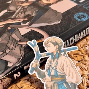 Peut inclure: Un autocollant représentant un personnage de style anime aux cheveux blonds, vêtu d'une tenue blanche et bleue, tenant un bâton bleu. L'autocollant est placé sur un manga avec le titre "GACHIAKUT" et le numéro "2".
