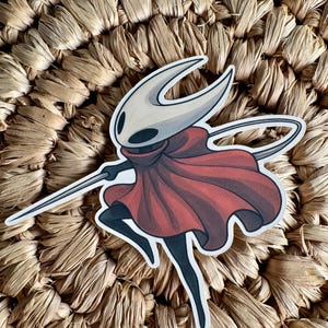 May include: A sticker featuring a character with a white mask, a red cape, and a silver blade. The character is in a dynamic pose, with one leg raised. The sticker is on a woven, beige background.