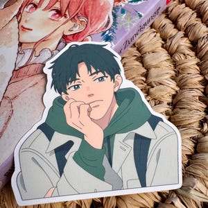 Può includere: Un adesivo con un'illustrazione in stile anime di una persona con i capelli verde scuro, che indossa una felpa con cappuccio verde sotto una giacca grigio chiaro. La persona appoggia il mento sulla mano. L'adesivo è su una superficie tessuta.
