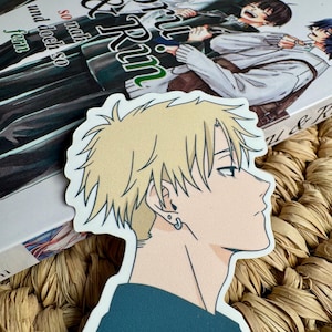 Peut inclure: Un autocollant représentant le profil d'un personnage d'anime blond portant un haut bleu et une boucle d'oreille. L'autocollant est posé sur un livre intitulé "Kaoru & Rin" avec un fond tissé.