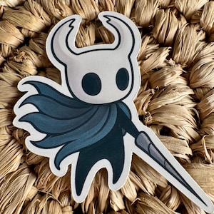May include: A sticker featuring a stylized character with a white head, large black eyes, and curved horns. The character wears a dark blue cape and holds a gray sword. The sticker is on a woven, textured background.