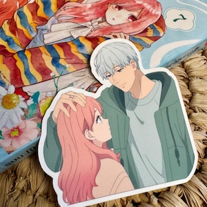 Può includere: Un adesivo con un'illustrazione in stile anime di una coppia. Il personaggio maschile ha i capelli argentati e indossa una felpa con cappuccio verde. Il personaggio femminile ha i capelli rosa e guarda in alto. L'adesivo è sopra un libro con un'illustrazione simile.