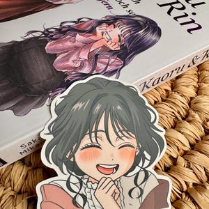 Peut inclure: Un autocollant représentant une illustration de style anime d'une jeune fille souriante aux cheveux foncés et aux joues roses. L'autocollant est placé devant un livre intitulé "Kaoru & Rin". La couverture du livre présente des illustrations et du texte de style anime.