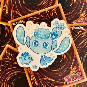 Pode incluir: Um autocolante azul claro com uma criatura de desenho animado com olhos grandes, um adorno de cabeça e múltiplos apêndices. O autocolante está em cima de vários cartões colecionáveis com um design flamejante e o texto "KONAMI" e "Yu-Gi-Oh! Trading Card Game".