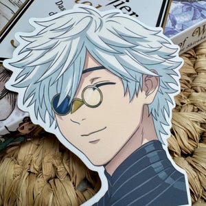 Könnte beinhalten: Ein gestanzter Aufkleber mit einem Anime-Charakter mit weißen Haaren und einem Monokel. Die Figur trägt ein dunkelblau gestreiftes Hemd. Der Aufkleber liegt auf einer gewebten Oberfläche, im Hintergrund ist ein Buch zu sehen.