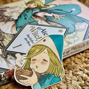 Könnte beinhalten: Ein Aufkleber im Anime-Stil mit einer Figur in einem türkis-weißen Hexenhut, die eine Schüssel hält. Der Aufkleber befindet sich auf einem Buch mit dem Titel "Ate Witch" und der Nummer "1". Der Hintergrund hat eine gewebte Textur.
