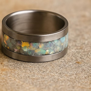 May include: A wide, brushed metal ring with a band of iridescent, multi-colored material. The ring's central band features a mosaic of colors including yellow, green, and blue. The ring is set against a neutral, textured background.