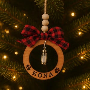 May include: A wooden Christmas ornament with a red and black plaid bow and the name "KONA" engraved on it. The ornament has paw prints and a small glass vial hanging in the center. The ornament is hanging from a string with wooden beads.