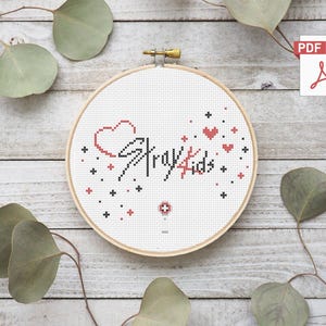 Peut inclure: Un cercle de broderie au point de croix avec le texte "Stray Kids" en fil noir et rouge. Le motif comprend un cœur et des étoiles. Le cercle en bois est posé sur un fond en bois.