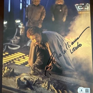 May include: A signed Star Wars movie still featuring Billy Dee Williams. The image shows Williams kneeling near a figure in a frozen state, with two alien figures in the background. The image is marked with the text "TM & © 1998 Lucasfilm Ltd. All Rights Reserved. Used under authorization."