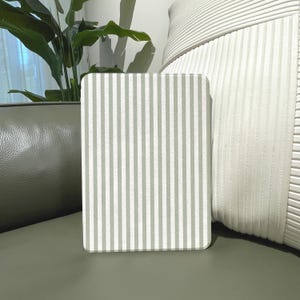 Peut inclure: Une planche décorative rectangulaire aux coins arrondis avec un motif à rayures verticales vert clair et blanc. La planche est debout, mettant en valeur le design. L'arrière-plan comprend une surface en cuir vert, une plante et un coussin blanc texturé.