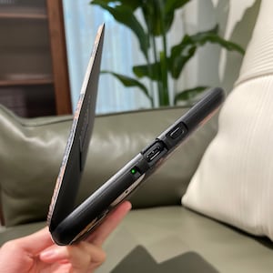 May include: Black phone case with a floral design and a matching flip cover. The case has a charging port and a power button. The flip cover is open, revealing the phone screen. The case is held in a person's hand.