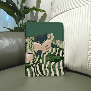 Puede incluir: Una funda rectangular para lector electrónico con fondo verde oscuro y una ilustración de un gato negro en la cama bajo una manta a rayas verdes y blancas, leyendo un libro con el texto "not smut" en la portada, junto a una taza de café.