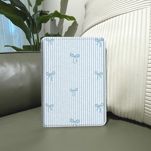 May include: A rectangular, light blue and white striped notebook cover with a pattern of small blue bows. The notebook is standing upright, with a green plant and a white pillow in the background.