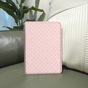 May include: A rectangular, light pink tablet case with a repeating pattern of small, darker pink dots. The case is standing upright on a green surface, with a plant and a white, textured pillow in the background.