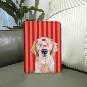 Pode incluir: Uma capa retangular com listras vermelhas e brancas, apresentando um golden retriever com beijos de batom. A capa, possivelmente para um tablet, repousa sobre uma superfície verde.