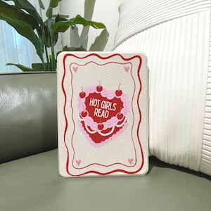 May include: A rectangular cream-colored decorative item with a red border and a heart-shaped cake design. The cake is pink and red, topped with cherries, and the text "HOT GIRLS READ". Small pink hearts are in each corner.