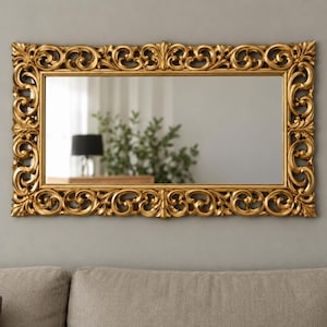 Pode incluir: Um espelho de parede retangular com uma moldura dourada metálica ornamentada com volutas intrincadas e entalhes em forma de folha. A superfície de vidro refletora é clara e lisa, emoldurada por uma borda decorativa espessa e texturizada.