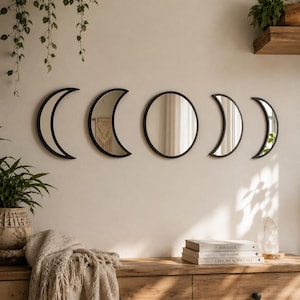 May include: A set of five wall-mounted mirrors featuring black frames arranged to represent the phases of the moon. The collection includes one circular mirror in the center, flanked by two crescent mirrors on each side, all with reflective glass.