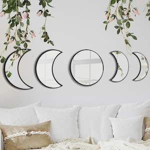 May include: A set of five wall-mounted mirrors with black frames, arranged to display the phases of the moon. The collection includes one circular mirror in the center, flanked by two crescent moon mirrors and two smaller crescent moon mirrors.