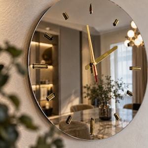 May include: A circular wall clock with a mirrored face and a thin silver frame. The clock features twelve gold-coloured cylindrical markers for the hours and two gold-coloured hands with a thin red accent line on the minute hand.