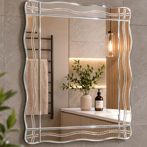 Puede incluir: Un espejo de pared rectangular con un marco decorativo de bordes de vidrio grabado ondulado y esquinas de vidrio transparente. La superficie del espejo refleja el interior de un baño con una encimera, una planta y una lámpara colgante.