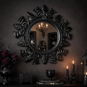 May include: A circular wall mirror featuring a matte black frame adorned with intricate, sculpted bat wing motifs and leaf-like details surrounding the glass. The frame has a dark, textured finish that creates a cohesive, monochromatic appearance.