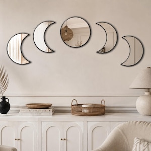 May include: A set of five wall-mounted mirrors arranged in a horizontal arc to represent the phases of the moon. The collection includes one central circular mirror flanked by four crescent-shaped mirrors, all featuring thin black frames.