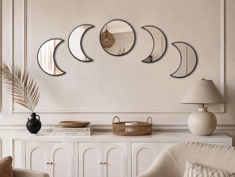 May include: A set of five wall-mounted mirrors arranged in a horizontal arc to represent the phases of the moon. The collection includes one central circular mirror flanked by four crescent-shaped mirrors, all featuring thin black frames.