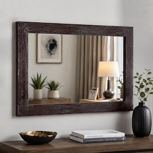 May include: A rectangular wall mirror featuring a dark brown frame with a textured, wood-grain finish. The mirror reflects a room interior containing a table lamp, two potted plants, and a framed picture, providing a clear, reflective surface.