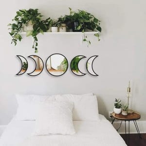 May include: A set of five wall-mounted mirrors arranged in a horizontal line to represent the phases of the moon. The set includes one circular mirror in the center flanked by four crescent-shaped mirrors with black frames, reflecting the room.