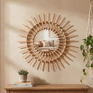 May include: A round, natural-toned rattan mirror with a sunburst design hangs on a beige wall. A small succulent in a gray pot sits on a wooden table below the mirror. A macrame plant hanger with a green plant is suspended from the ceiling.