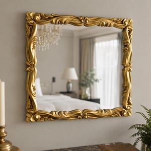 May include: A square wall mirror featuring a thick, ornate gold frame with intricate, flowing scrollwork and carved detailing. The reflective glass surface displays a clear view of a bedroom interior, including a lamp and a chandelier.