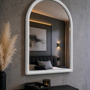 Puede incluir: Un espejo de pared arqueado con un marco blanco brillante que presenta sutiles detalles acanalados horizontales en los lados inferiores. El espejo tiene una parte superior redondeada y una base plana, mostrando un reflejo de un dormitorio.