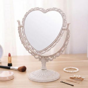 May include: A white, heart-shaped vanity mirror with an ornate, scrolling design. The mirror is double-sided and sits on a decorative base. Makeup items, including lipstick and a brush, are visible on the wooden surface.